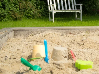 Adelheidshof Sandkasten Sandkasten mit buntem Spielzeug im Vordergrund und einem weißen Holzbank im Grünen im Hintergrund.