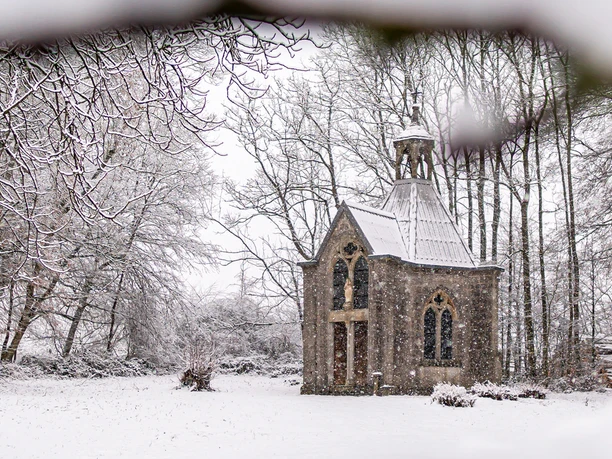 Eine kleine Kapelle steht malerisch im winterlichen Wald, umgeben von schneebedeckten Bäumen.