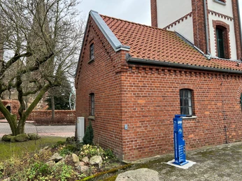 Radstation Bücken Ziegelgebäude mit rotem Dach und blauer Radladestation, das sich an einer Pflasterstraße befindet.