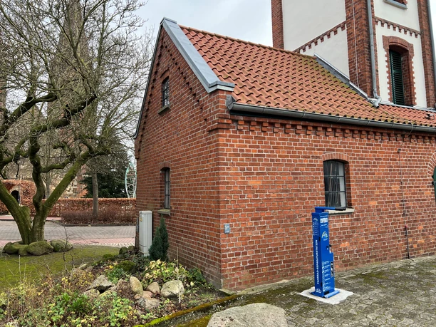 Radstation Bücken Ziegelgebäude mit rotem Dach und blauer Radladestation, das sich an einer Pflasterstraße befindet.