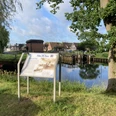 Fehn-Tour_Schild_mit_Erklärung_an_der_Schleuse_Rhauderfehn_IMG_5759.JPG Informationsschild am Ufer eines Kanals mit Blick auf eine Schleuse und Häuser im Hintergrund unter blauem Himmel