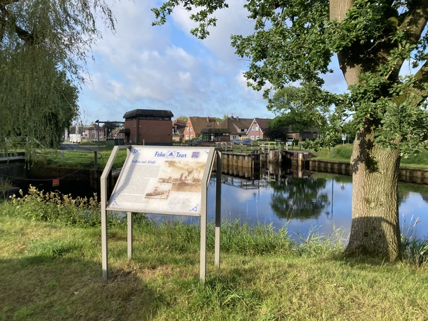 Fehn-Tour_Schild_mit_Erklärung_an_der_Schleuse_Rhauderfehn_IMG_5759.JPG Informationsschild am Ufer eines Kanals mit Blick auf eine Schleuse und Häuser im Hintergrund unter blauem Himmel