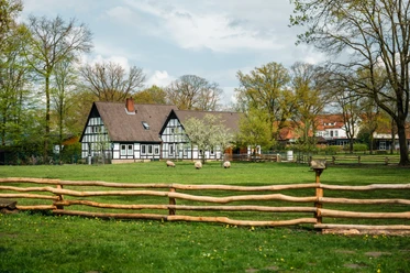 Fachwerkgebäude mit dunklem Dach, umgeben von grüner Wiese, Bäumen und Schafen hinter Holzzaun.