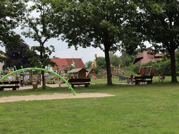 Spielplatz mit Klettergerüst, Schaukeln und Rasenflächen umgeben von großen Bäumen, ideal für Familien.