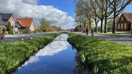 Wiekentour (Radthemenroute) - Gemeinde Rhauderfehn, 1. Südwieke Kanal mit Himmelspiegelung in Rhauderfehn, beidseitig Straßen mit Radfahrern und Backsteinhäusern.