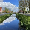 Wiekentour (Radthemenroute) - Gemeinde Rhauderfehn, 1. Südwieke Kanal mit Himmelspiegelung in Rhauderfehn, beidseitig Straßen mit Radfahrern und Backsteinhäusern.