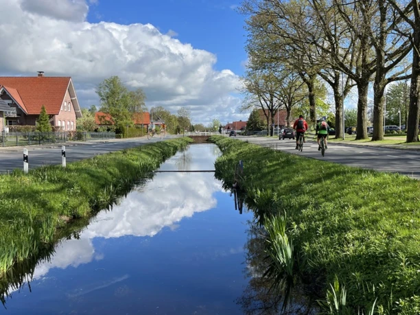Wiekentour (Radthemenroute) - Gemeinde Rhauderfehn, 1. Südwieke Kanal mit Himmelspiegelung in Rhauderfehn, beidseitig Straßen mit Radfahrern und Backsteinhäusern.