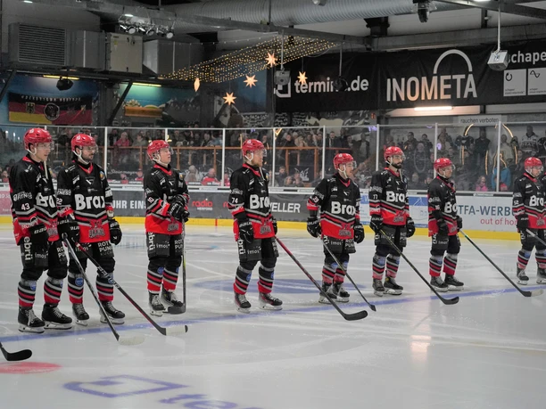 Eishockeyspieler in roten Helmen stehen auf dem Eis, bereit für das nächste Spiel. Zuschauertribüne im Hintergrund.