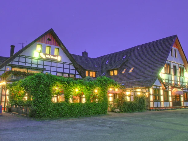 Fachwerkhotel mit beleuchteter Fassade, vor dörflicher Kulisse in der Dämmerung, begrüntes Vorhaus.