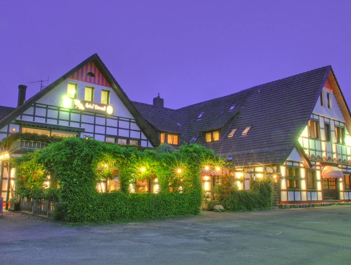 Fachwerkhotel mit beleuchteter Fassade, vor dörflicher Kulisse in der Dämmerung, begrüntes Vorhaus.