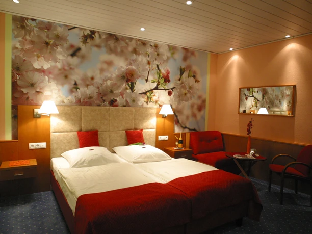 Doppelzimmer mit zwei Einzelbetten, blumiger Akzentwand, rotem Stuhl und Tisch unter warmem Licht.