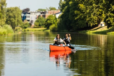 Zwei Personen paddeln entspannt in einem roten Kanu auf einem ruhigen Fluss, umgeben von Natur und Bäumen.
