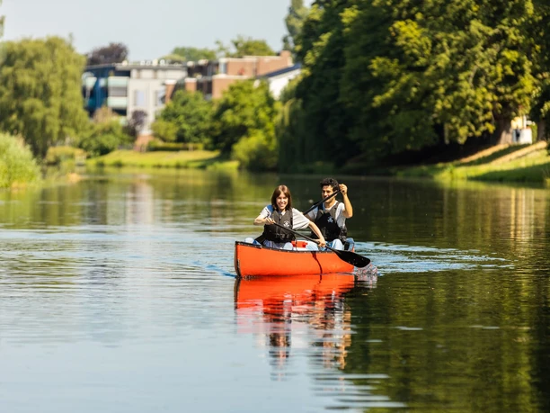 Zwei Personen paddeln entspannt in einem roten Kanu auf einem ruhigen Fluss, umgeben von Natur und Bäumen.