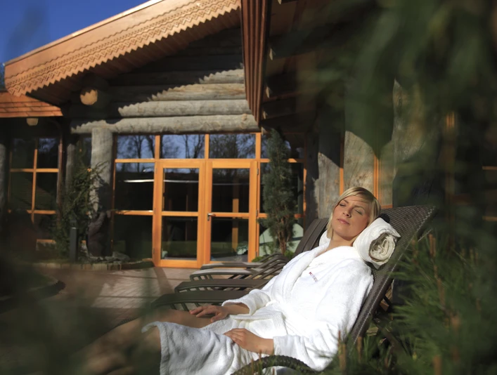 h2o-haus-19.JPG Eine Frau in einem weißen Bademantel entspannt auf einer Liege vor einem Holzgebäude mit großen Fenstern.