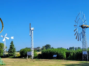 Deutsches Windkraftmuseum Verschiedene historische Windkraftanlagen auf grüner Wiese, umgeben von Bäumen und blauem Himmel.