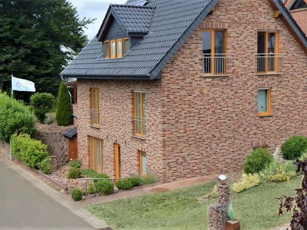 Ferienwohnung in rotem Backsteingebäude mit schrägem Dach, gepflegtem Garten und Weg an ruhiger Straße.