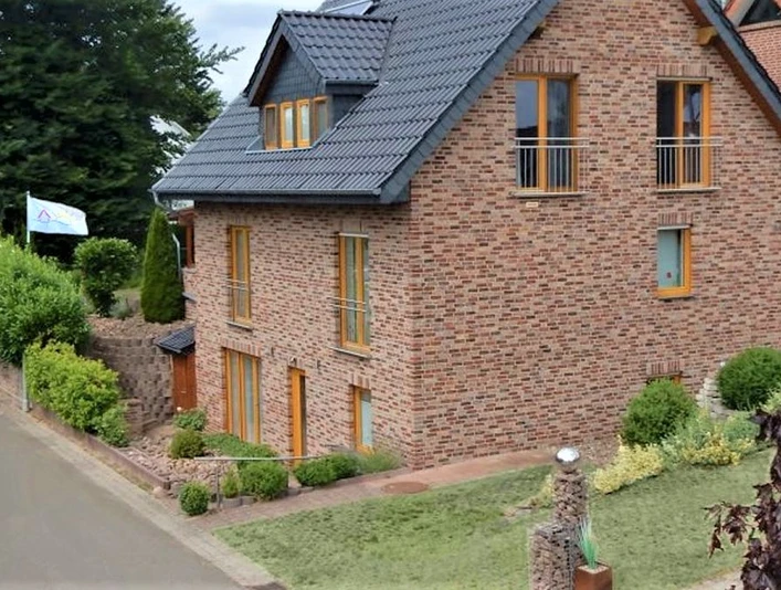 Ferienwohnung in rotem Backsteingebäude mit schrägem Dach, gepflegtem Garten und Weg an ruhiger Straße.
