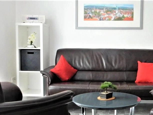 Wohnzimmer einer Ferienwohnung mit schwarzem Ledersofa, roten Kissen, Couchtisch und Wandbild.