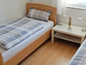 Zweibettzimmer mit heller Holzmöblierung, zwei Betten, weißem Nachttisch und großen Fenstern.