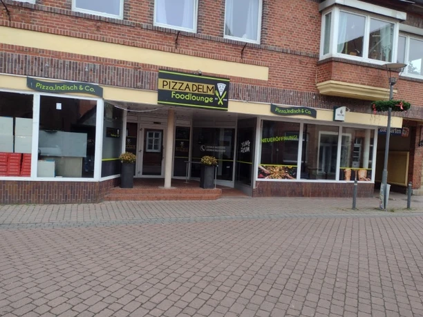 Pizza Delm 1.jpg Eingang der Pizzeria Pizza Delm mit gelbem Schild und großer Fensterfront in rotem Backsteingebäude.