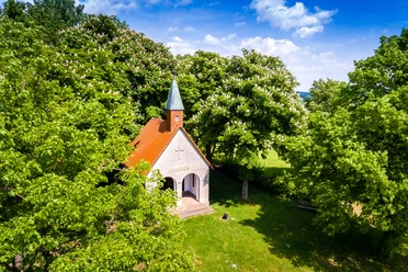 Altenheerse-Himmelsbergkapelle-4.jpg Eine kleine Kapelle mit rotem Dach und grünem Turm umgeben von blühenden Kastanienbäumen.