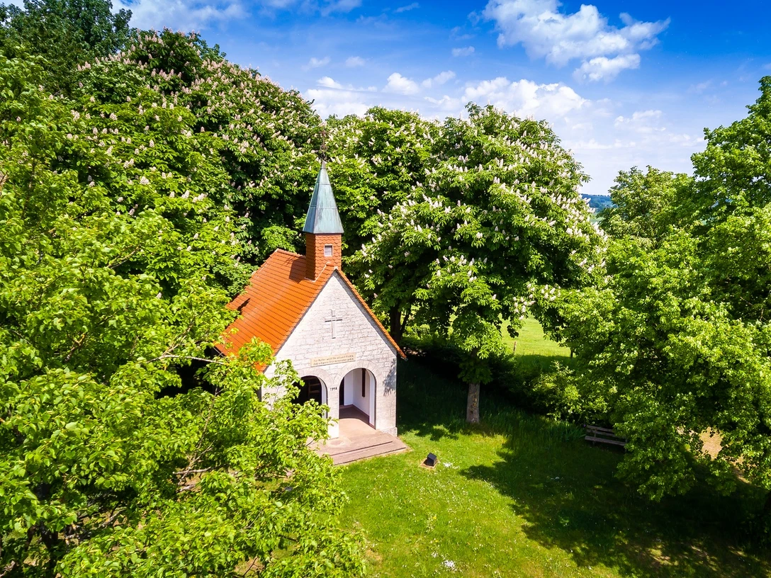 Altenheerse-Himmelsbergkapelle-4.jpg Eine kleine Kapelle mit rotem Dach und grünem Turm umgeben von blühenden Kastanienbäumen.
