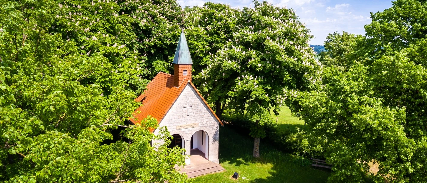 Altenheerse-Himmelsbergkapelle-4.jpg Eine kleine Kapelle mit rotem Dach und grünem Turm umgeben von blühenden Kastanienbäumen.