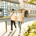 herford-altstadt-teutoburger-wald-tourismus-h-tornow-003-jpg.jpg Ein Paar spaziert an Fachwerkhäusern in einer sonnigen Altstadt, umgeben von grüner Vegetation.