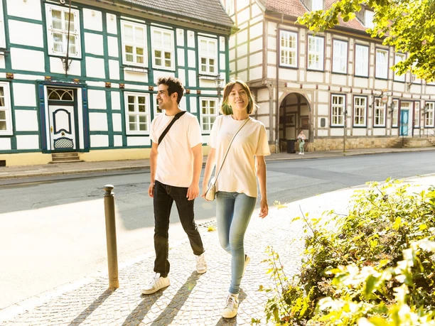 herford-altstadt-teutoburger-wald-tourismus-h-tornow-003-jpg.jpg Ein Paar spaziert an Fachwerkhäusern in einer sonnigen Altstadt, umgeben von grüner Vegetation.