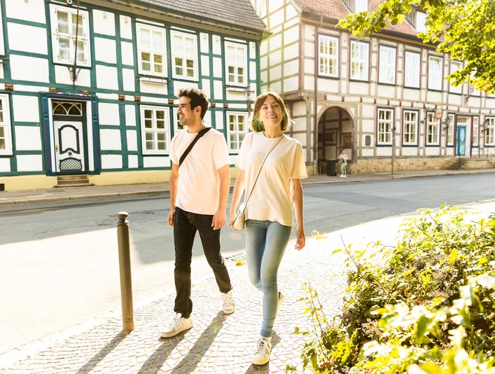 herford-altstadt-teutoburger-wald-tourismus-h-tornow-003-jpg.jpg Ein Paar spaziert an Fachwerkhäusern in einer sonnigen Altstadt, umgeben von grüner Vegetation.