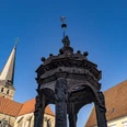 Herford_Herbst_2020_017.jpg Steinerner Brunnen vor historischer Kirche mit Langschiff, flankiert von traditionellen Gebäuden.
