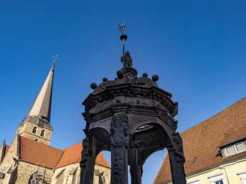 Herford_Herbst_2020_017.jpg Steinerner Brunnen vor historischer Kirche mit Langschiff, flankiert von traditionellen Gebäuden.
