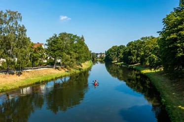 herford-kanu-teutoburger-wald-tourismus-h-tornow-001-cc-by-sa-jpg.jpg Zwei Personen paddeln in einem roten Kanu auf einem ruhigen Fluss, gesäumt von grünen Bäumen.