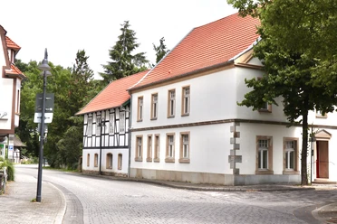 Straßenszene mit Fachwerkhaus und roten Dächern, umgeben von Bäumen und ruhiger Atmosphäre.