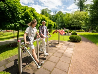 Besucher genießen die Nutzung von Outdoor-Fitnessgeräten inmitten einer grünen Parklandschaft.