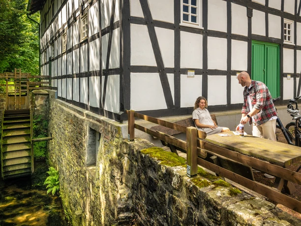Eine historische Wassermühle mit Fachwerk, umgeben von Natur, mit einem Paar an einem Holztisch.