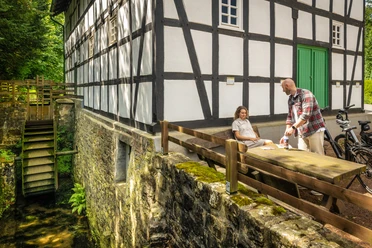 Gutswassermühle Bad Holzhausen Eine historische Wassermühle mit Fachwerk, umgeben von Natur, mit einem Paar an einem Holztisch.
