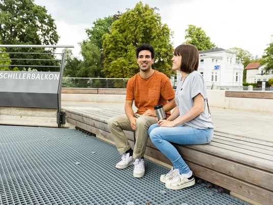 herford-promenade-teutoburger-wald-tourismus-h-tornow-001-jpg.jpg Zwei Menschen sitzen entspannt auf einer Bank mit Holzplanken am Schillerbalkon in Herford.