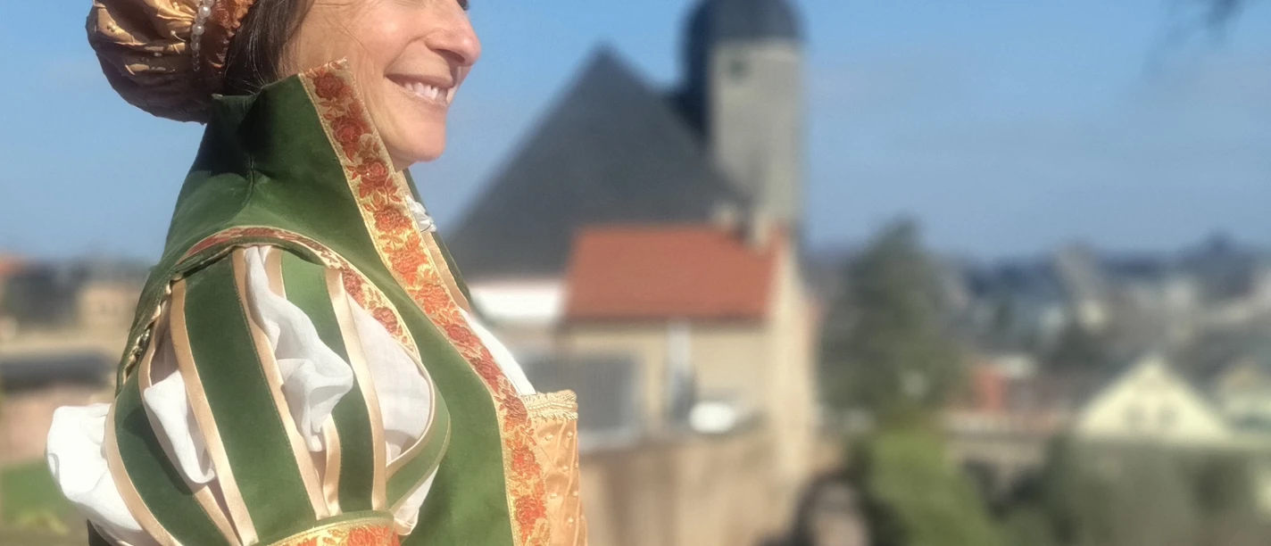 Die skandalöse Herzogin Schloss Rochlitz Frau in Renaissancekleid lächelt auf Schlossmauer mit Turm und städtischer Kulisse im Hintergrund.