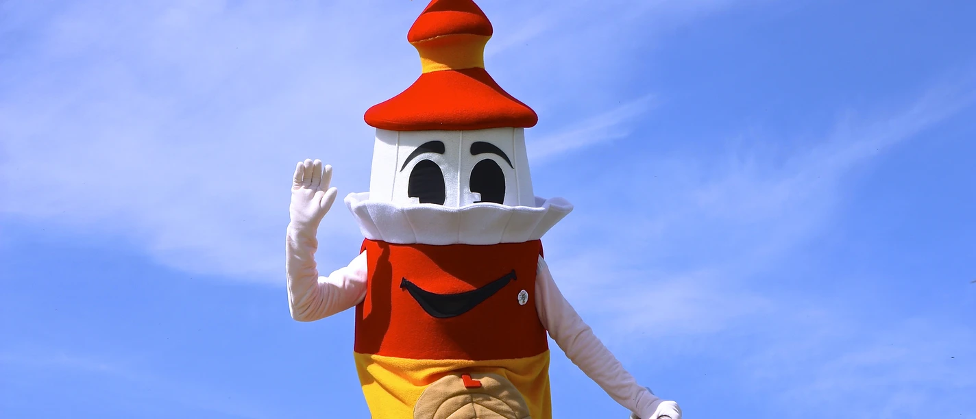 A mascot in a lighthouse costume with a red tower roof waves cheerfully against a blue sky.