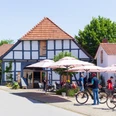 Radlerpause in Levern.jpg Eine Gruppe von Radfahrern pausiert vor einem Fachwerkhaus mit schattigen Sonnenschirmen in Levern.