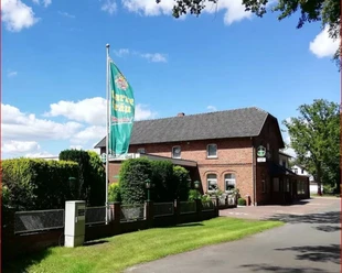 Ilweder_Hof.jpg Ein Backsteinhaus in ländlicher Umgebung mit einer großen Fahne und gepflegtem Garten.