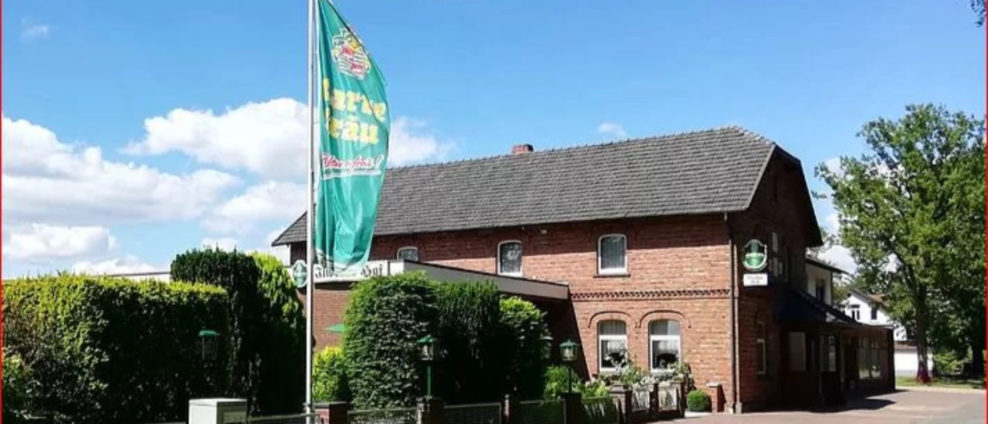 Ilweder_Hof.jpg Ein Backsteinhaus in ländlicher Umgebung mit einer großen Fahne und gepflegtem Garten.