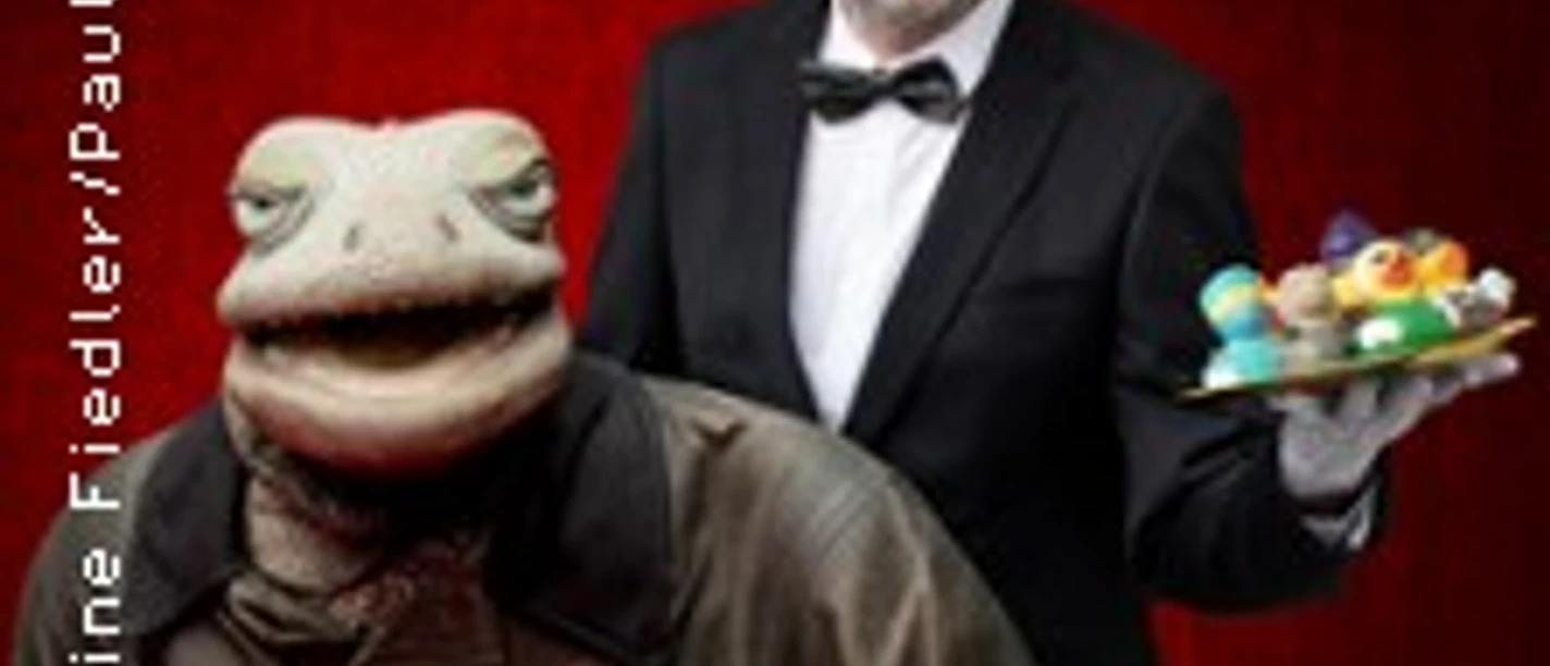 Ein als Kellner gekleideter Mann posiert mit einer großen Marionette eines alten Mannes vor rotem Hintergrund.