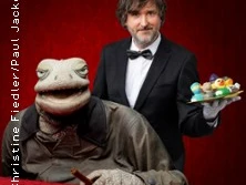 Ein als Kellner gekleideter Mann posiert mit einer großen Marionette eines alten Mannes vor rotem Hintergrund.