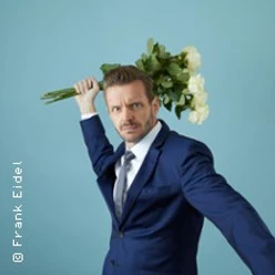 Ein Mann in blauem Anzug hält fröhlich einen Blumenstrauß über der Schulter vor blauem Hintergrund.