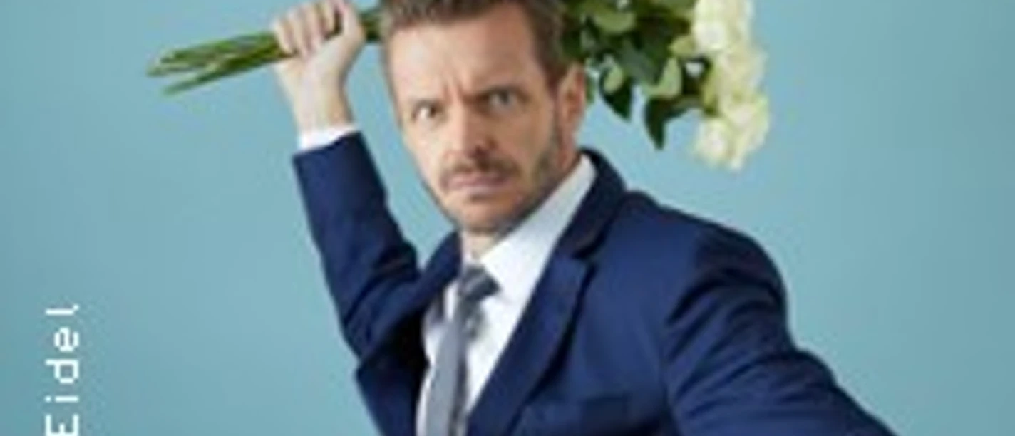 Ein Mann in blauem Anzug hält fröhlich einen Blumenstrauß über der Schulter vor blauem Hintergrund.