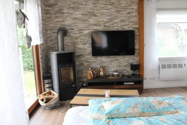 Een lichte woonkamer met houtkachel, flatscreentelevisie en grote ramen met uitzicht op de tuin.