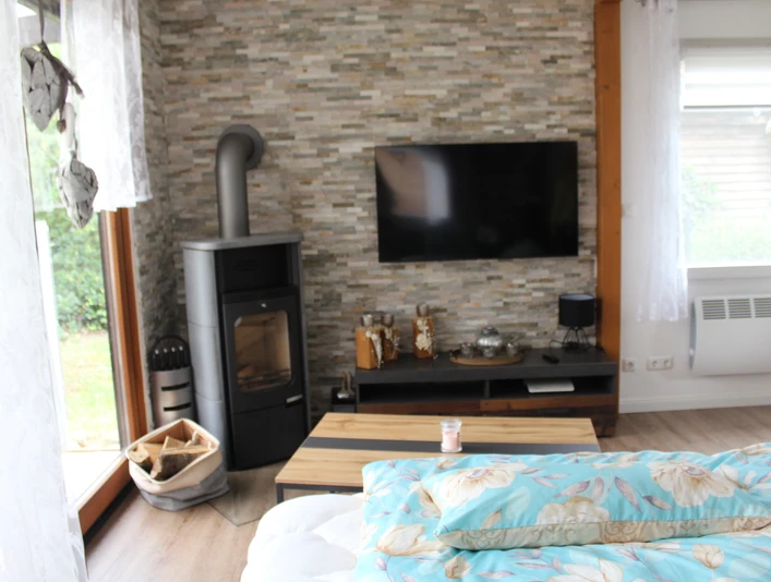 Ein helles Wohnzimmer mit Kaminofen, Flachbildfernseher und großen Fenstern, Blick in den Garten.