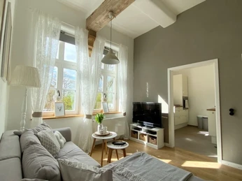 Helles Wohnzimmer mit Holzdecke, Sofa, Fernseher, Beistelltisch und Blick in die angrenzende Küche.